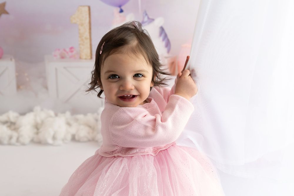 Young child wearing pink tulle dress smiles and waves in decorated white room with party decorations.