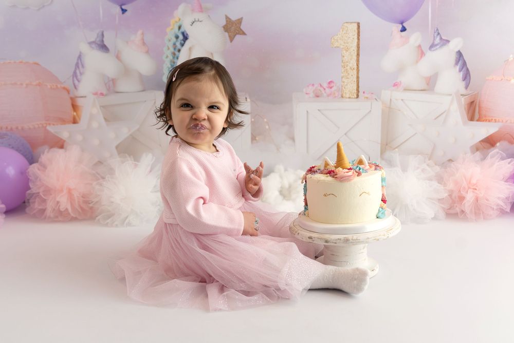 First birthday cake setup with unicorn theme featuring pink tulle, balloons, and white backdrop for celebration photoshoot.