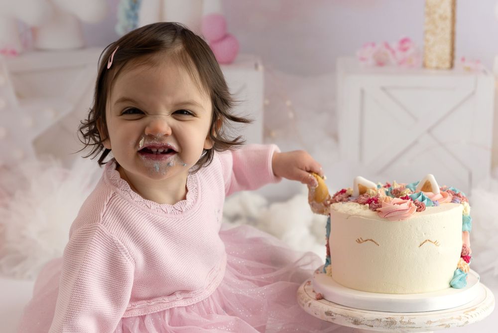 A happy toddler in a pink sweater sits next to a decorated white cake with sprinkles and unicorn-style decorations.