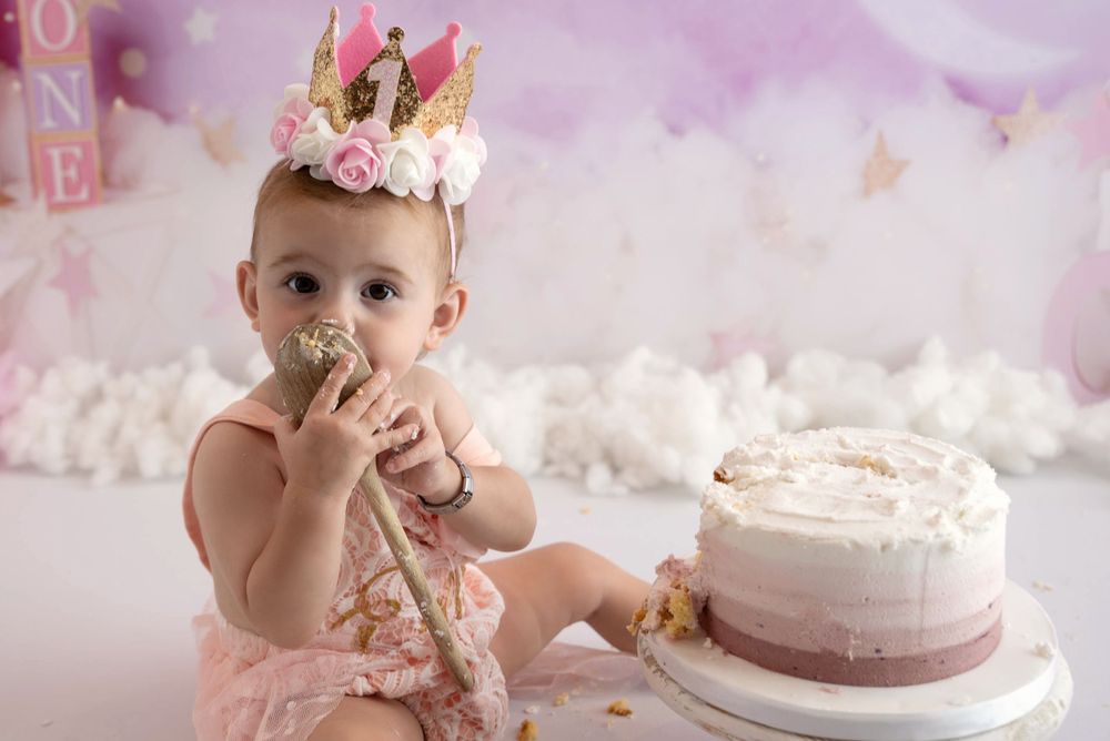 Baby in pink dress and crown smashes birthday cake during first birthday cake smash photoshoot with purple background.