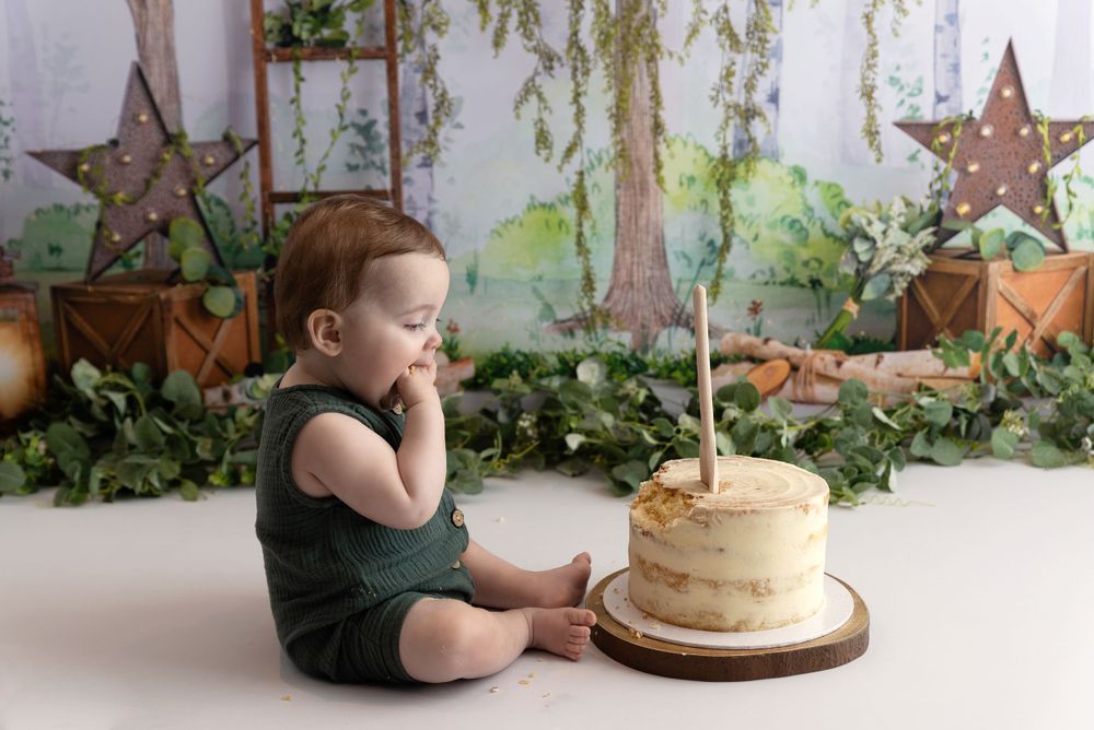 Baby in dark outfit sits thoughtfully next to simple white birthday cake with single candle against garden backdrop.