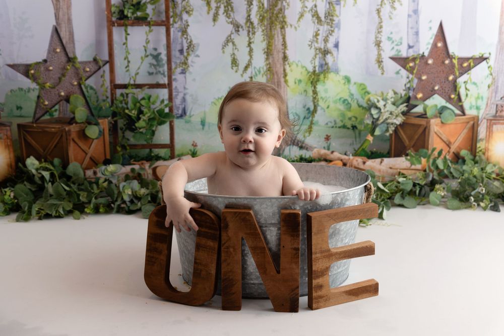 Baby sits inside rustic wooden ONE sign on white backdrop with decorative greenery and stars.