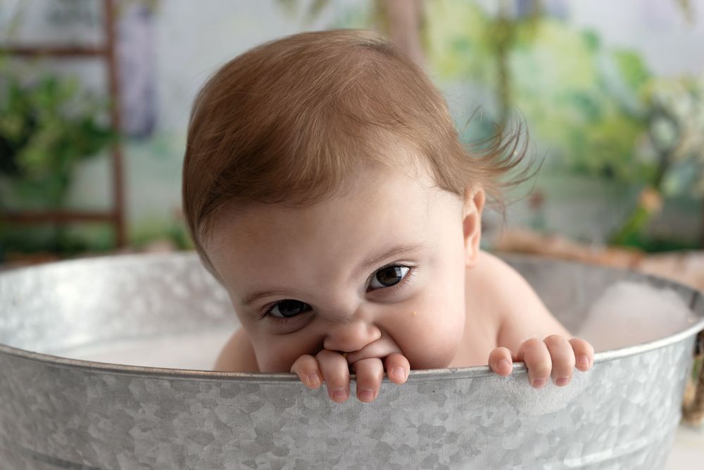 A small baby peers over the edge of a silver metal tub with just their eyes visible above the rim.