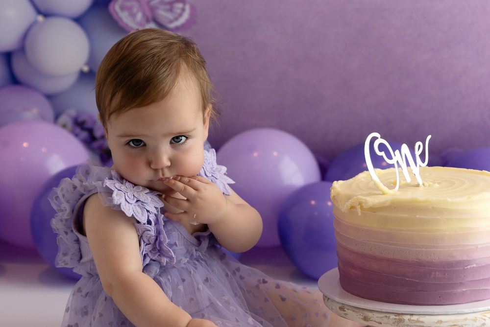 Purple ombre birthday cake with balloons and a toddler in a lavender tulle dress sitting beside it.