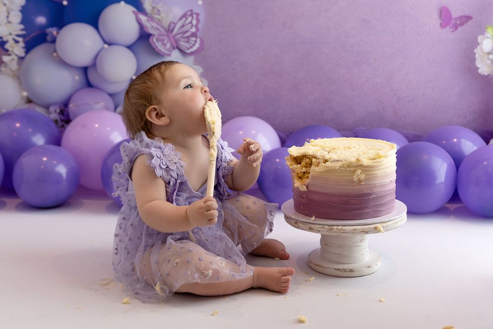 A small child in a purple dress sits on the floor with a cake and purple balloon decorations during a cake smash photo session.
