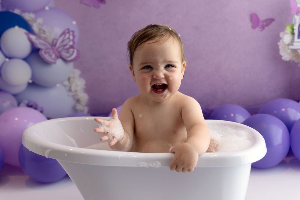 Happy baby sitting in white bathtub with purple and blue balloon decorations and butterfly accents in background.