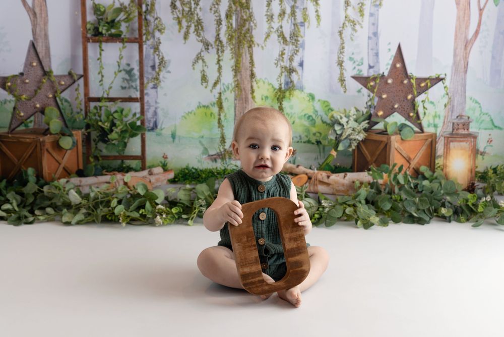 A small child in denim overalls sits on a white floor with rustic decor and greenery in the background.