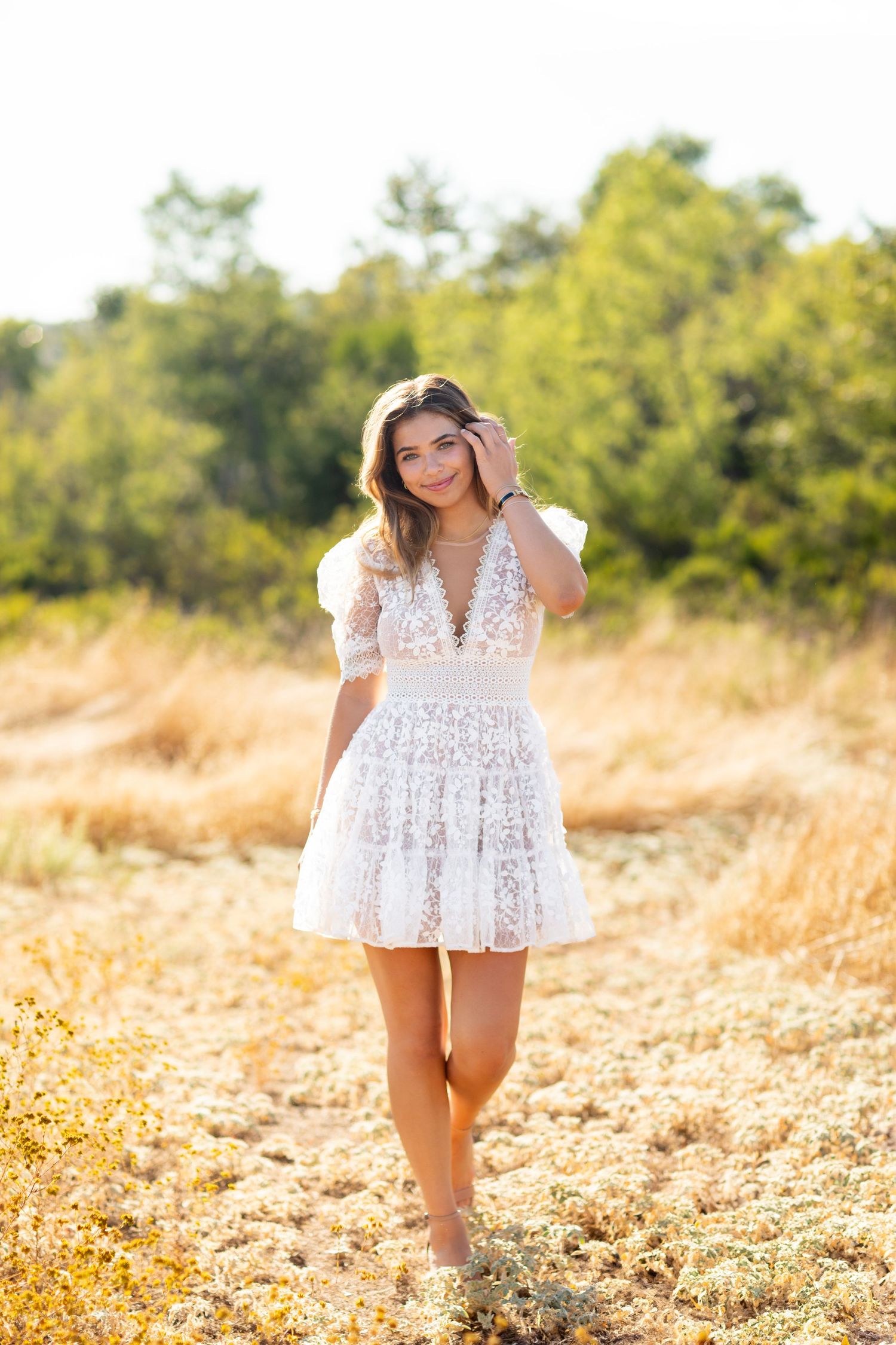 A high school senior wearing a white lace dress elegantly stands out against a golden field and green trees in the warm sunlight.