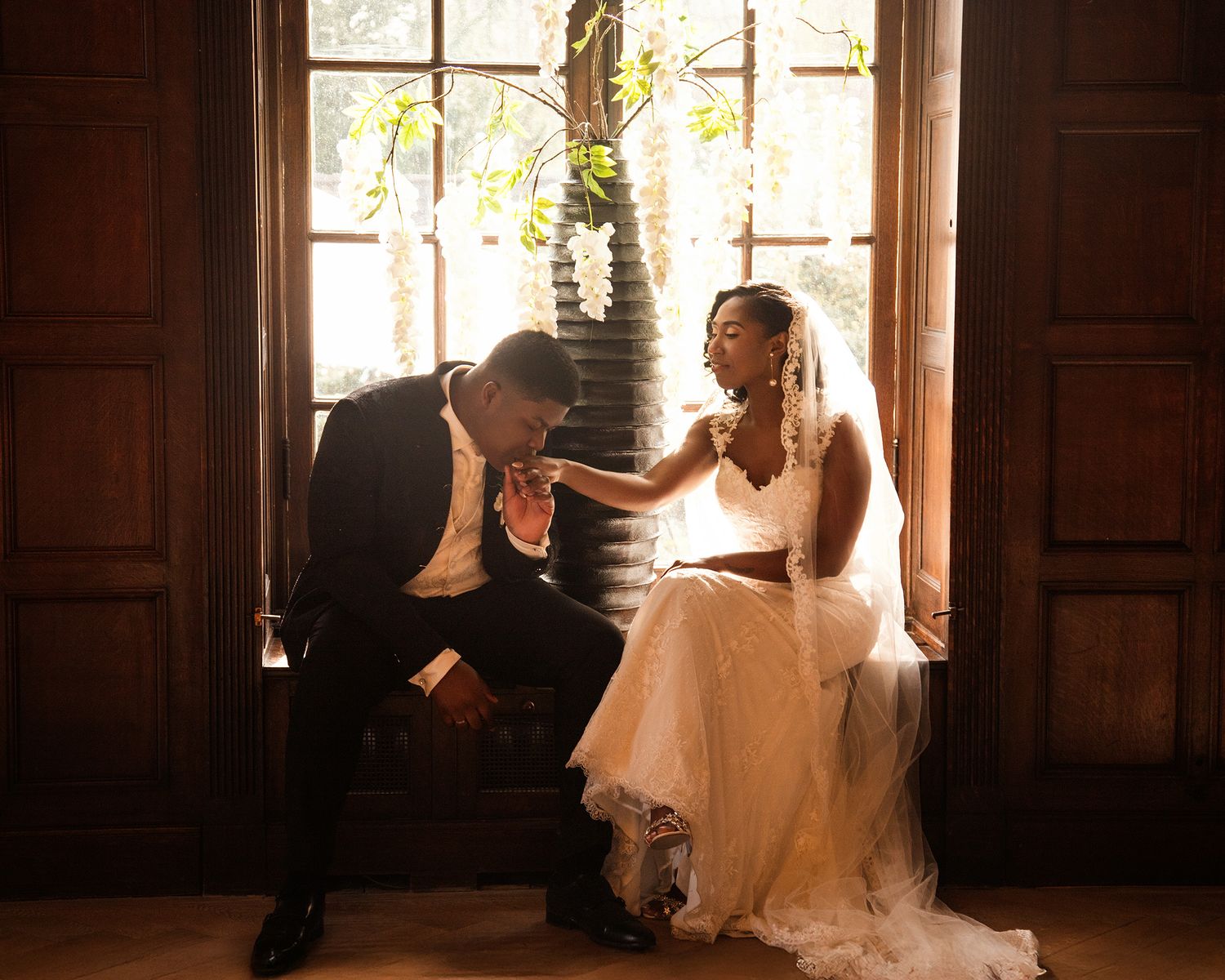 A couple in wedding attire sits together by a window in warm lighting, with the groom kissing the bride's hand.