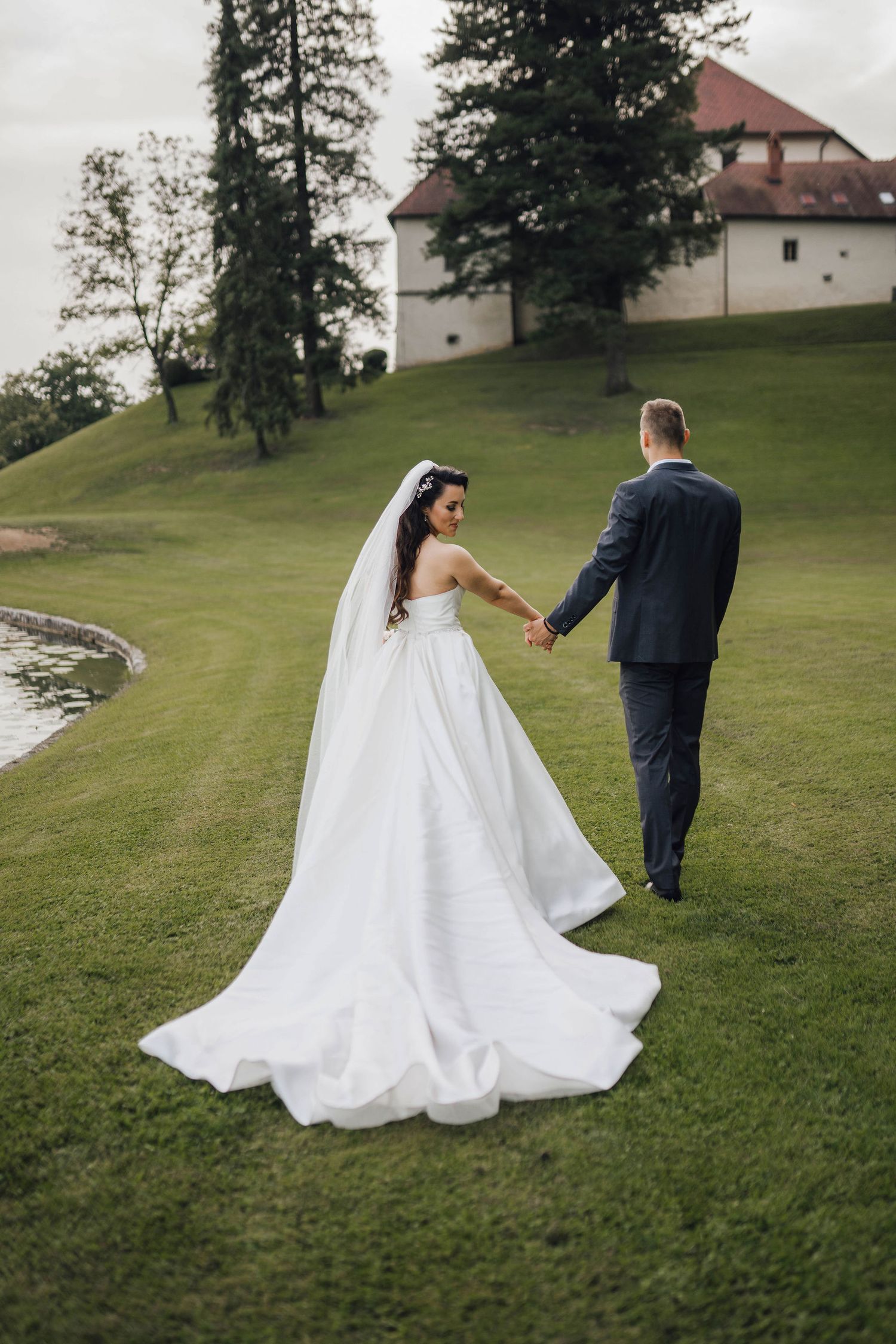 Newlywed couple walking hand in hand on grassy hillside near historic building.