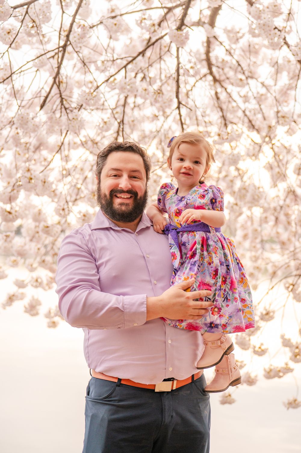 A touching spring portrait under blooming cherry blossom trees with soft pink petals creating a dreamy backdrop.
