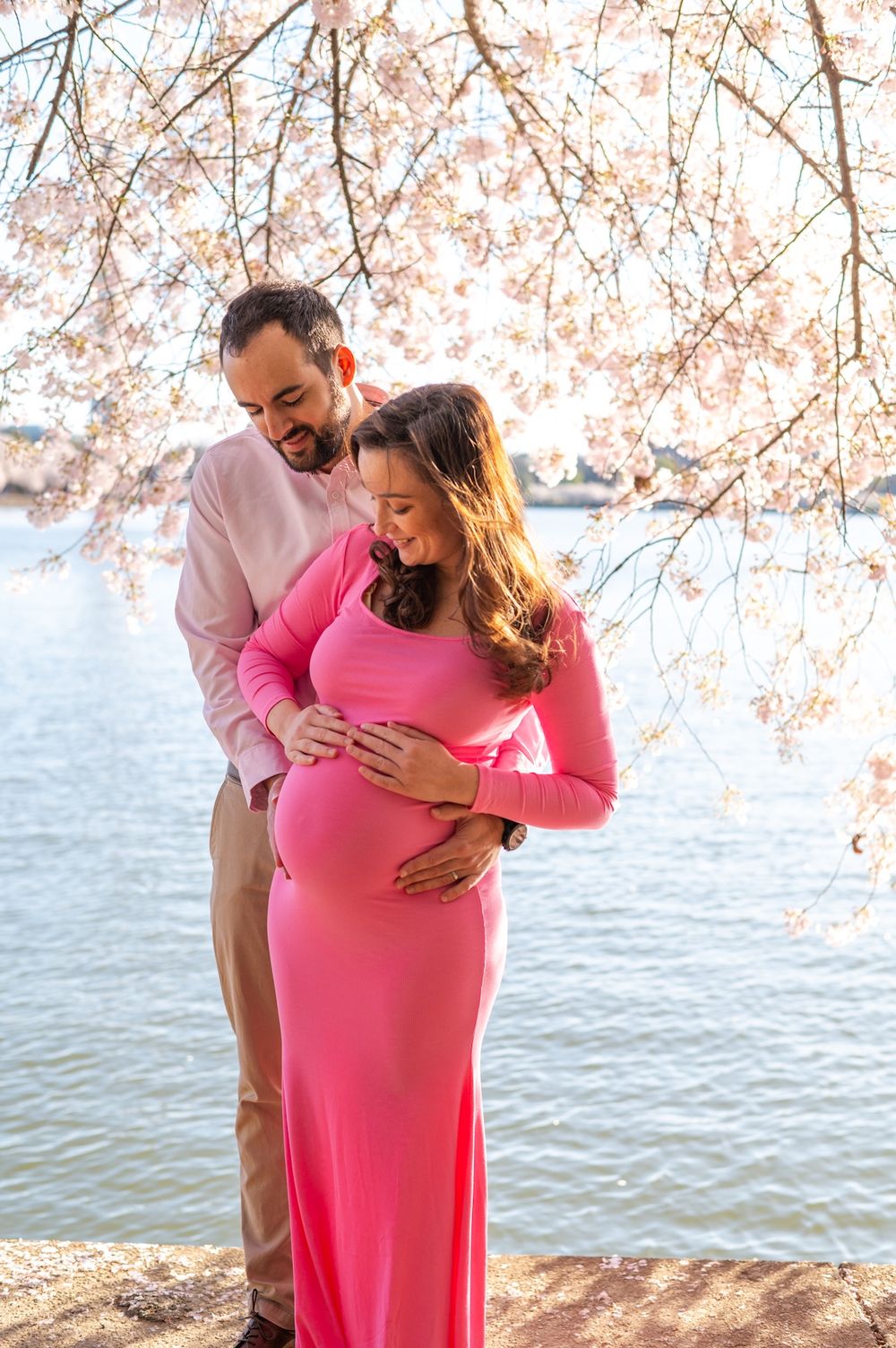 A couple embraces with her baby bump underneath cherry blossoms by a tranquil waterfront during a romantic spring photoshoot.
