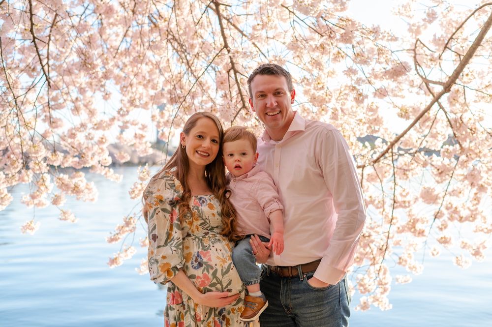 A family poses together under blooming cherry blossom trees near a waterfront during springtime.