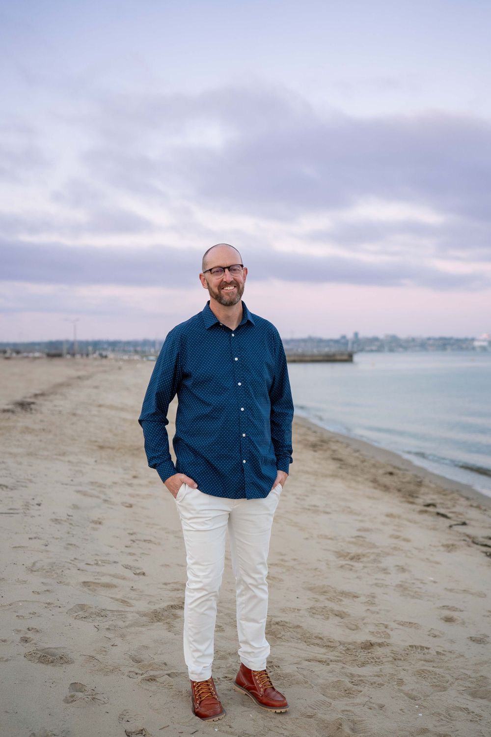 Man in blue button-down shirt and white pants stands on beach at dusk.