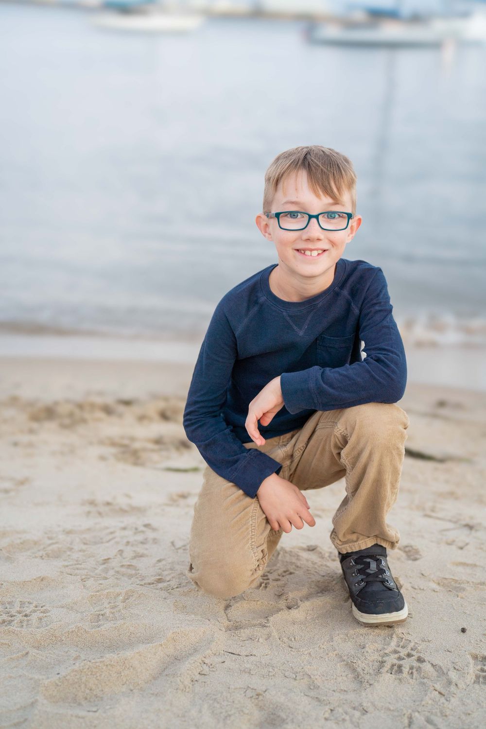 Young child in navy sweater crouches on sandy beach wearing blue glasses.
