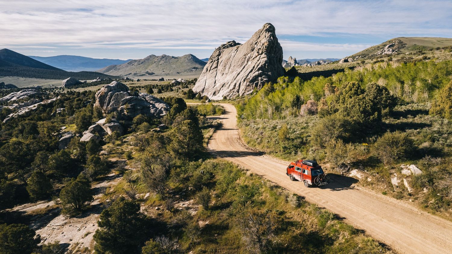 A bright red campervan drives along a winding dirt road through rugged mountain terrain with dramatic rock formations.