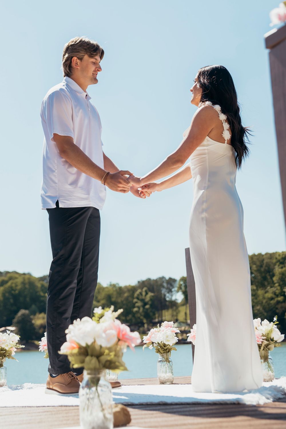 Couple holding hands at outdoor waterfront wedding ceremony with pink flowers and blue sky.