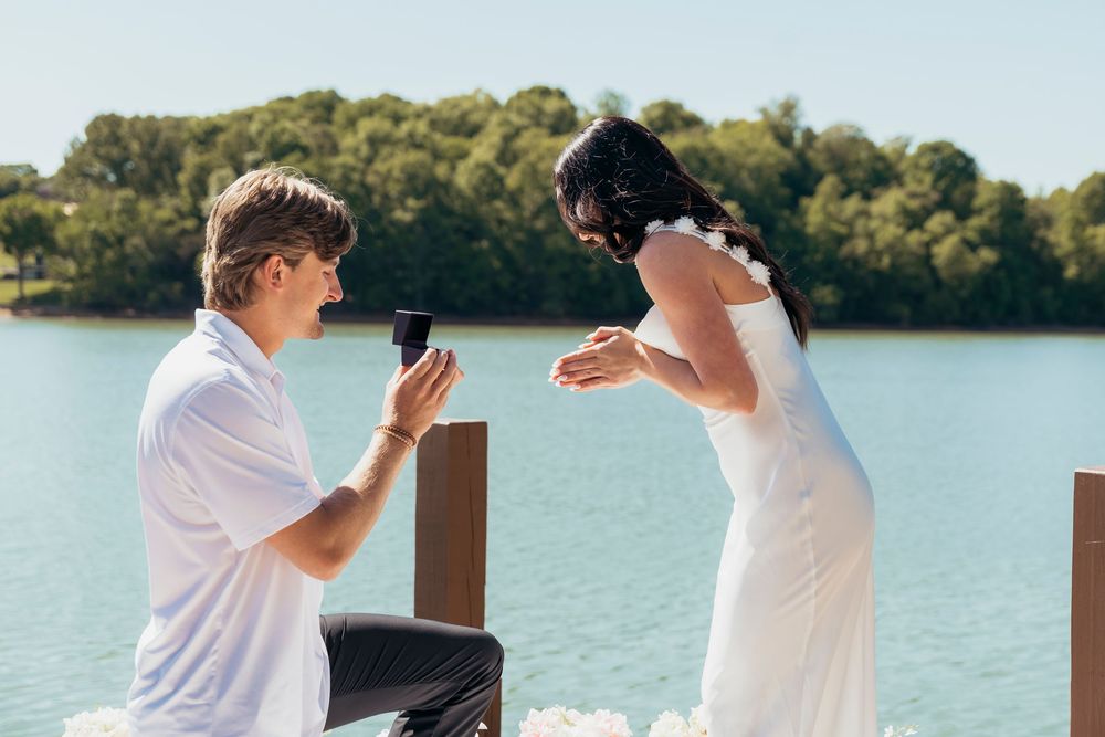 Man kneeling on a dock proposing to a woman in a white dress by a serene blue lake on a sunny day.