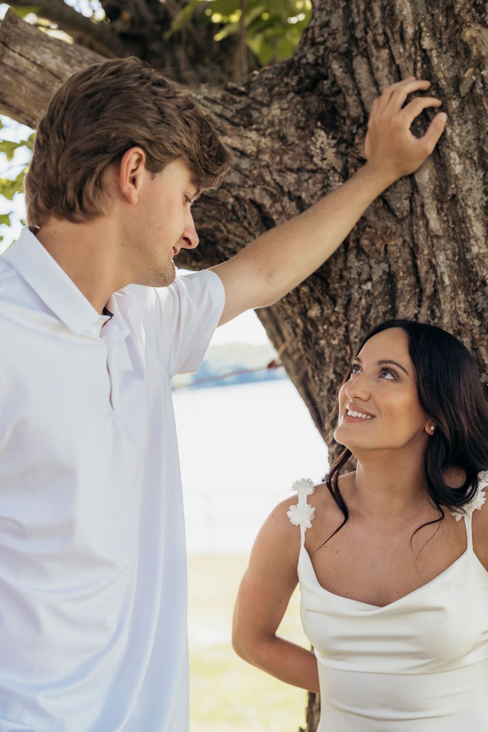 Couple in white attire sharing a romantic moment under a tropical tree on a sunny beach.