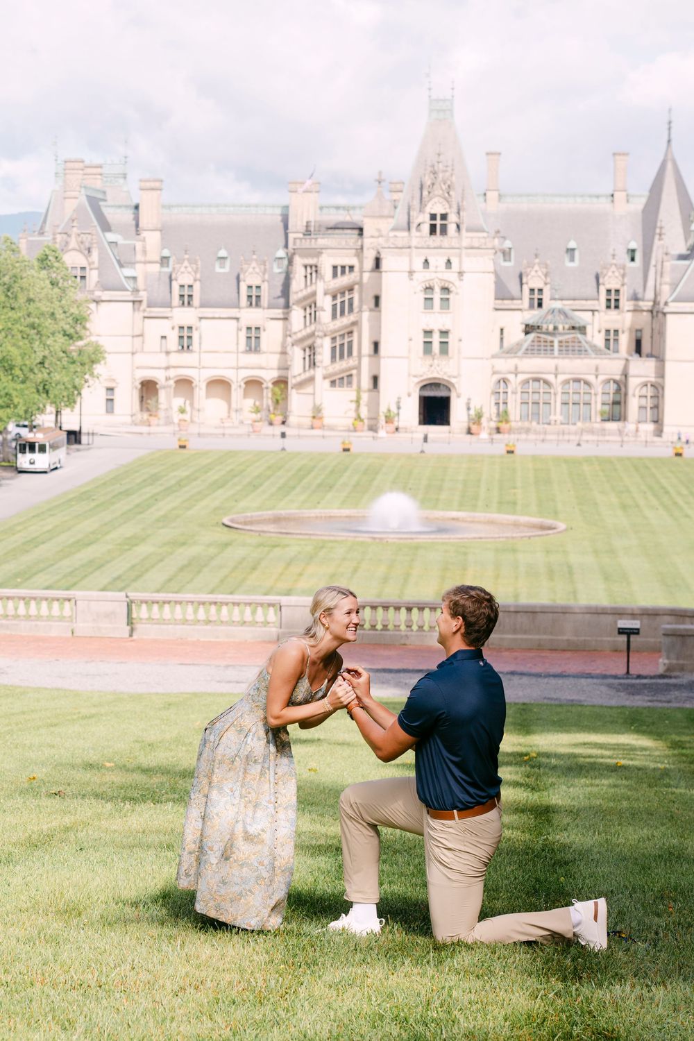 Man proposes on one knee to woman in floral dress on lawn in front of grand Biltmore Estate mansion.