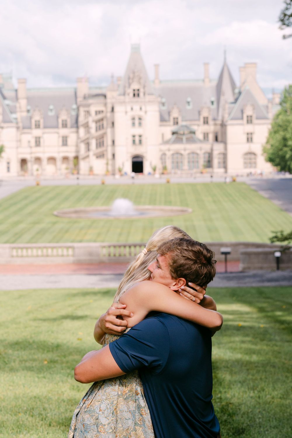 Couple embracing in front of the grand Biltmore Estate mansion with lush green lawn and fountain backdrop.