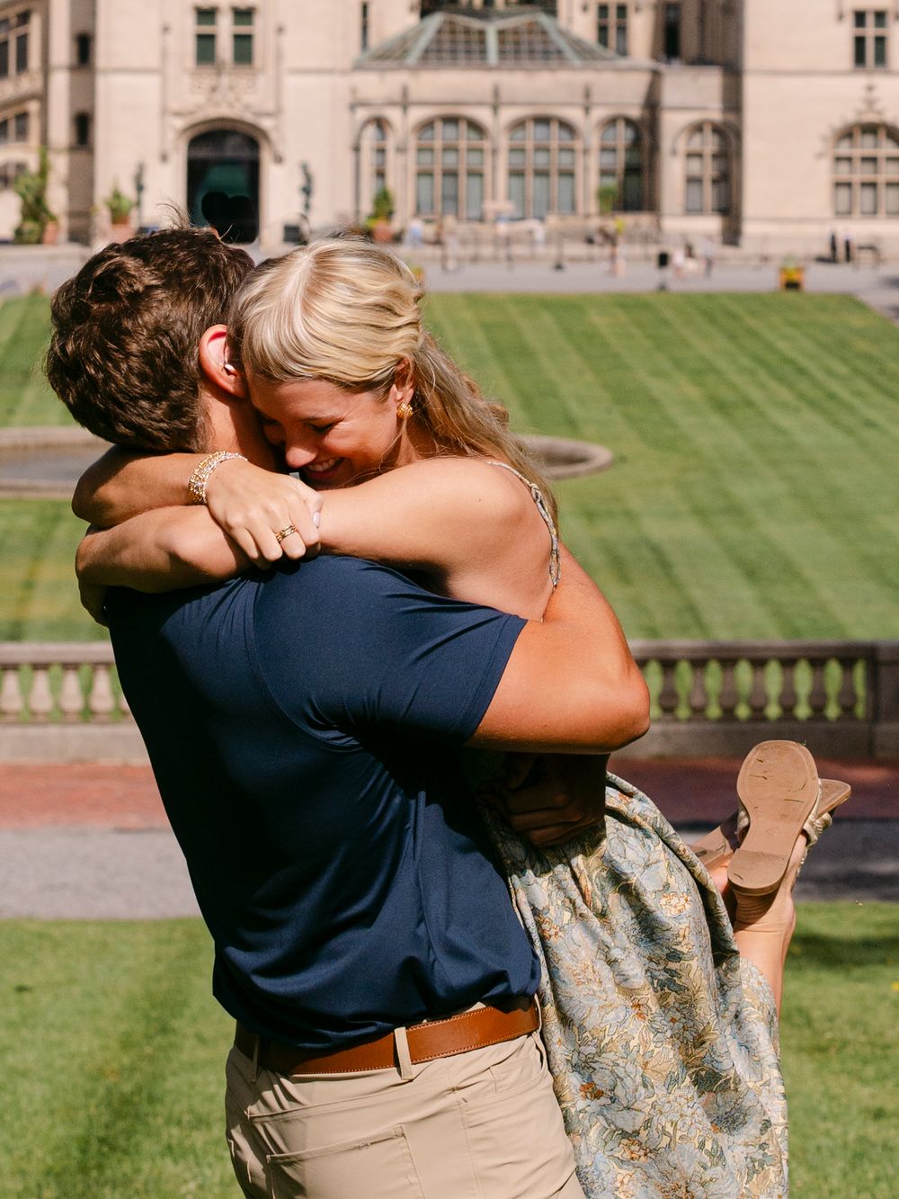 Couple embracing in front of a grand estate during an outdoor marriage proposal on a sunny day.