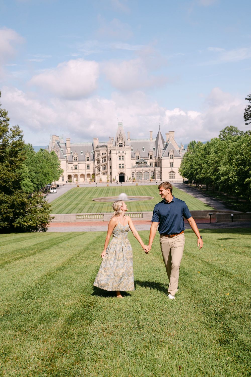 Couple holding hands walking on green lawn with Biltmore Estate mansion in background on sunny day.