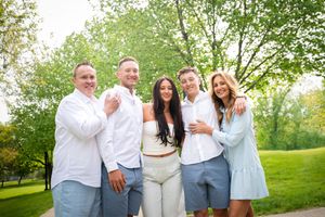 Family of five smiling outdoors in coordinated white and blue outfits surrounded by lush green trees.