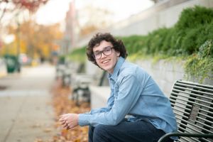 A casual portrait of someone sitting on a bench wearing glasses and a denim shirt on an autumn day.