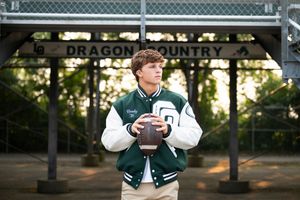 Teen boy in green varsity jacket holds football in front of Dragon Country sign for senior portraits.