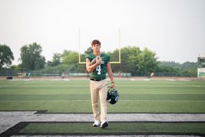 Teen football player in green jersey number 4 poses on turf field holding helmet near goal post.