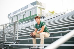 A young football player in green uniform sits on bleachers holding a football at an outdoor stadium.