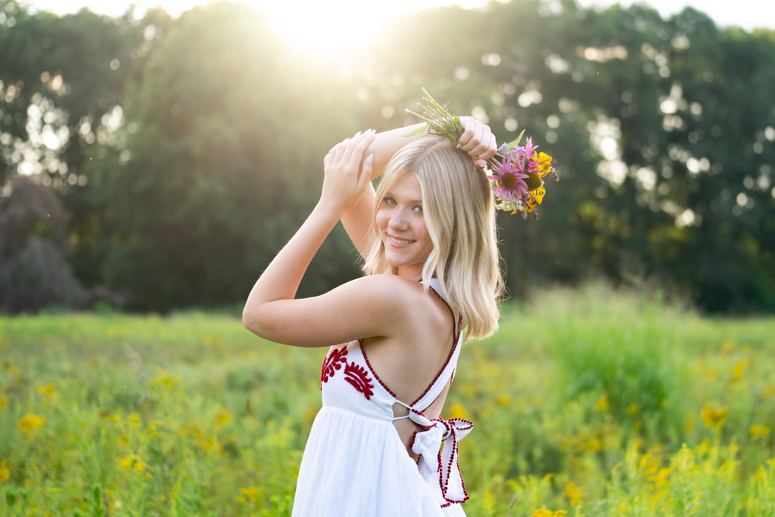 Smiling blonde woman wearing a white dress and floral crown standing in a sunny green meadow with wildflowers.