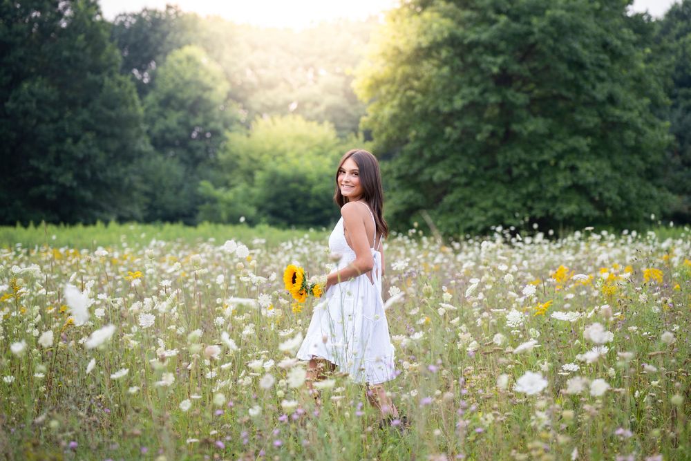 A white sundress stands out in a summer meadow filled with white and yellow wildflowers at golden hour.
