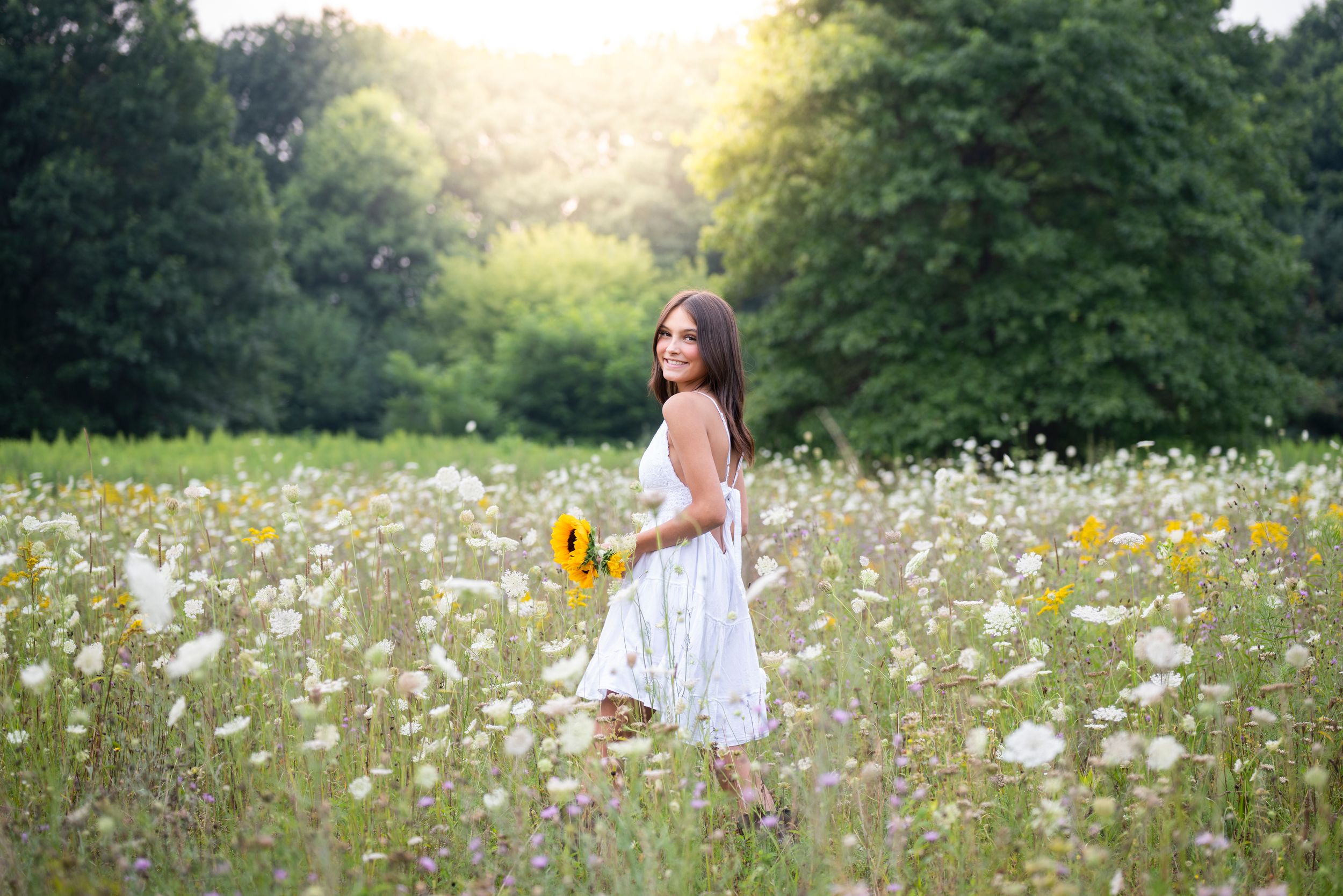 A white sundress stands out in a summer meadow filled with white and yellow wildflowers at golden hour.