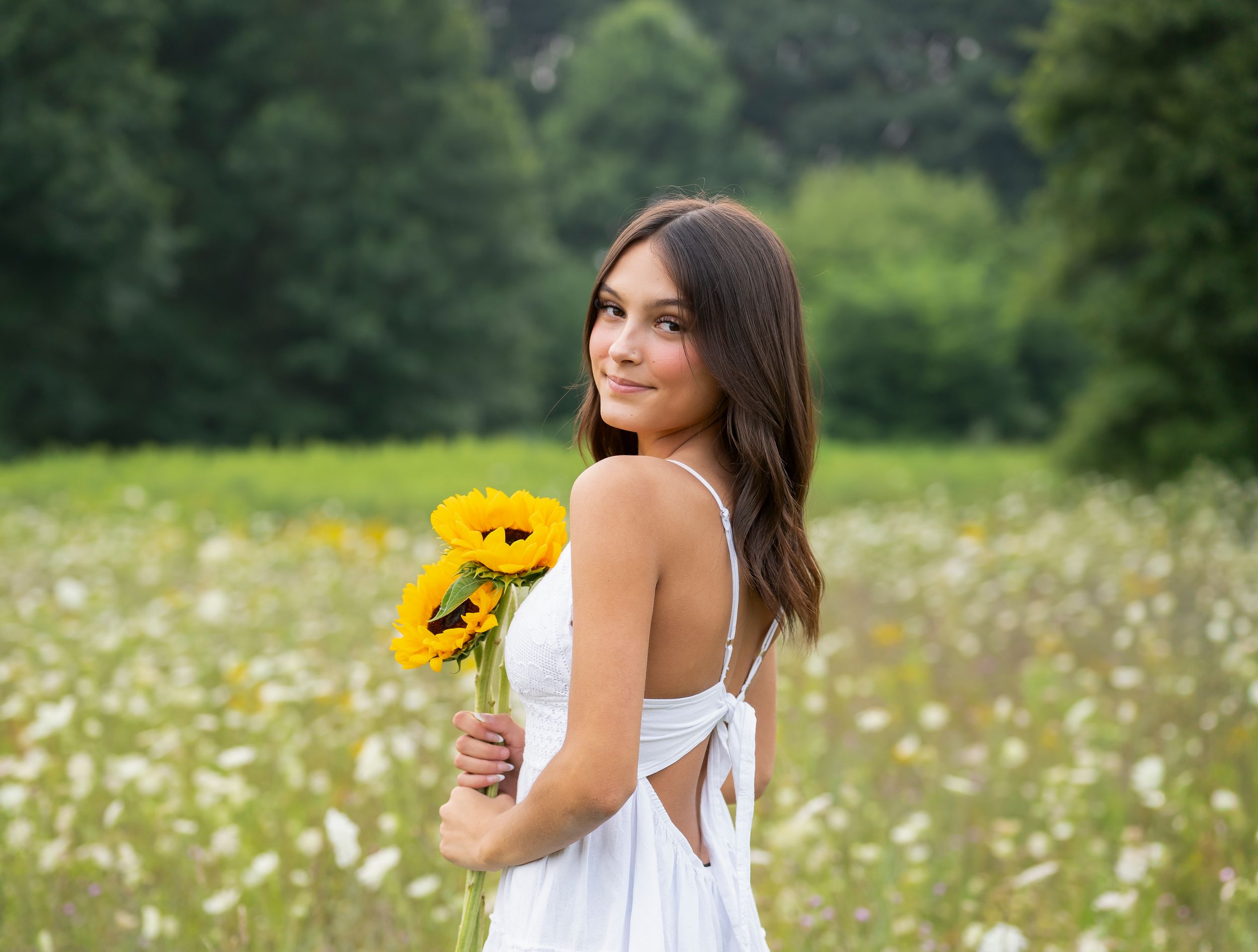 Young woman in white dress holding sunflowers in a blooming meadow with lush green trees in the background.