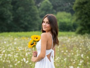 Young woman in white dress holding sunflowers in a blooming meadow with lush green trees in the background.