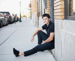 Young man in black outfit sitting against brick building on urban sidewalk, smiling confidently.