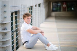 Student in white polo and gray pants sitting against wall in school hallway.