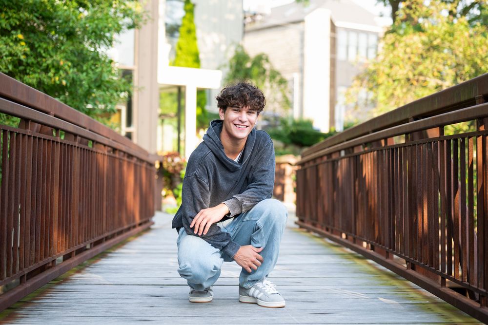Smiling teenage boy crouching on a wooden bridge outdoors, wearing a gray hoodie and jeans.