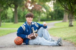 Student athlete relaxing on path with basketball and guitar in varsity jacket and jeans on sunny day.