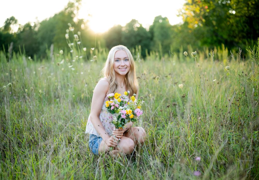 Smiling blonde girl kneeling in a sunny meadow holding a colorful wildflower bouquet surrounded by tall green grass.