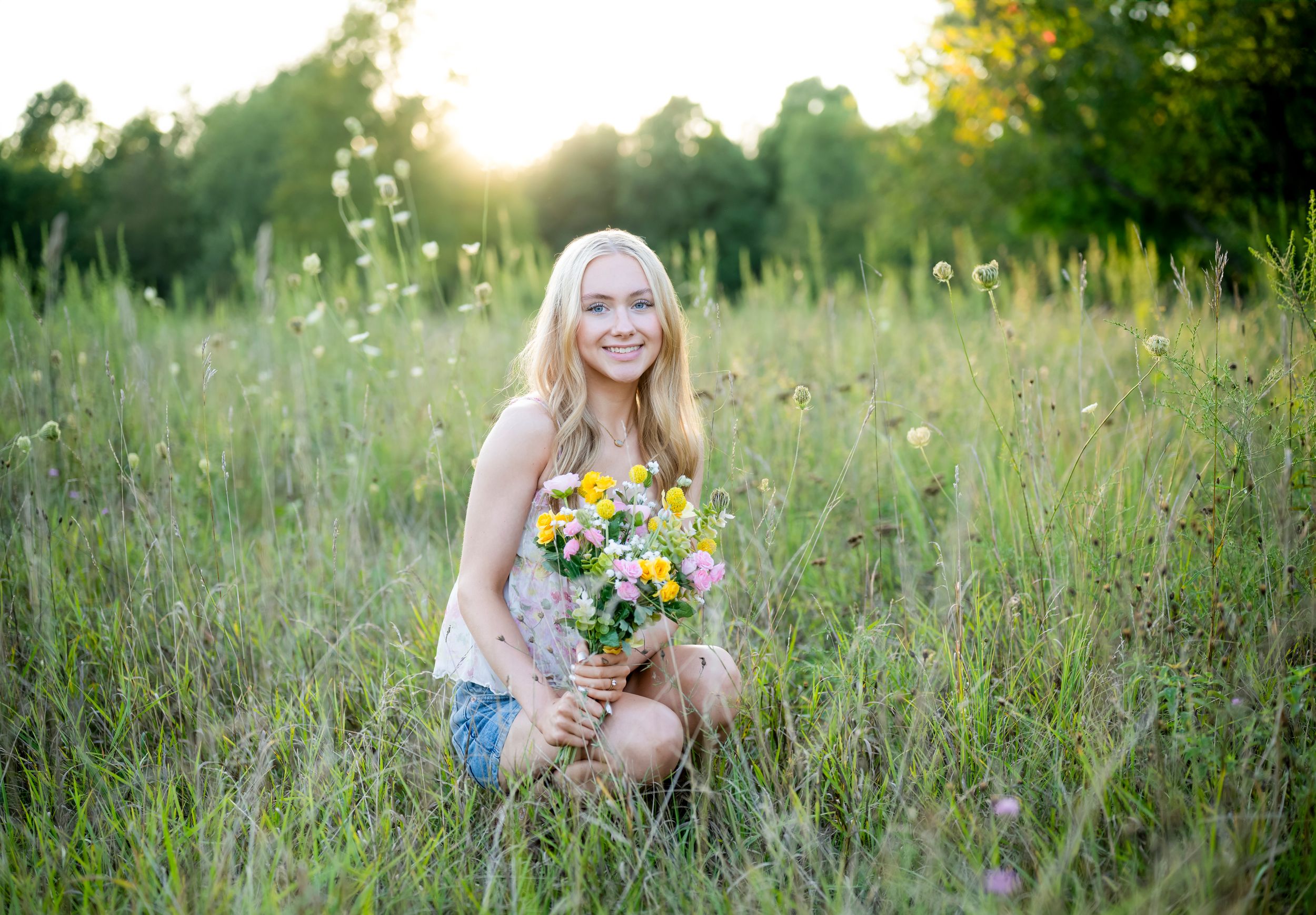 Smiling blonde girl kneeling in a sunny meadow holding a colorful wildflower bouquet surrounded by tall green grass.
