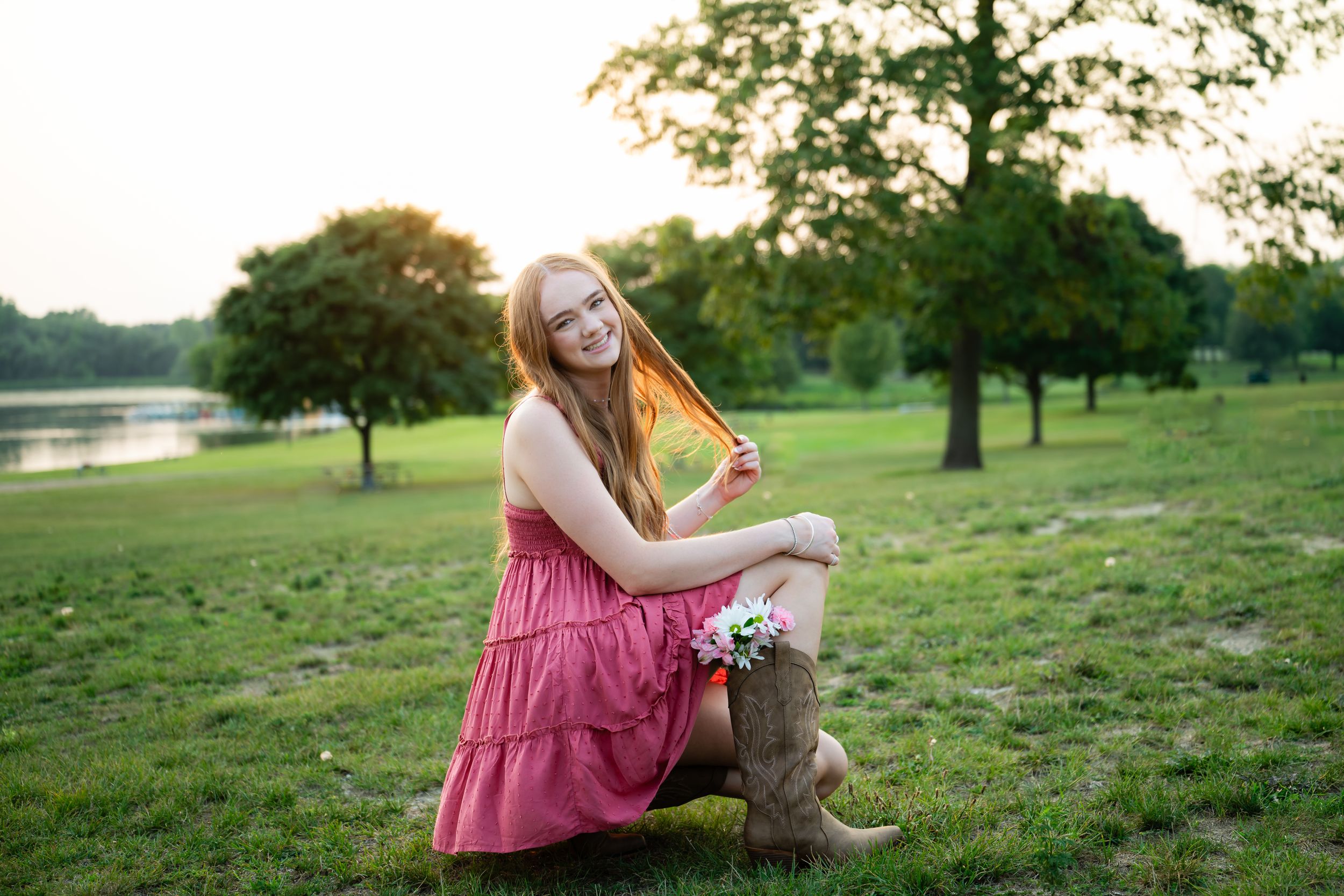 Young redhead woman in pink dress and cowboy boots sits on stump in park near lake at golden hour.