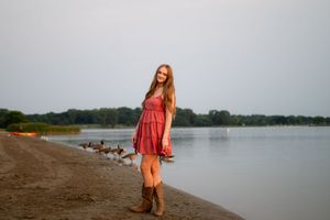 Young woman in red dress and cowboy boots smiling on a lakeside beach with geese in the background.