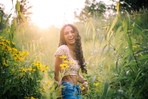 Person posing in summer attire among tall grass and wildflowers at golden hour.