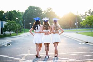 Three graduates in white dresses and caps walk arm-in-arm into the sunset.