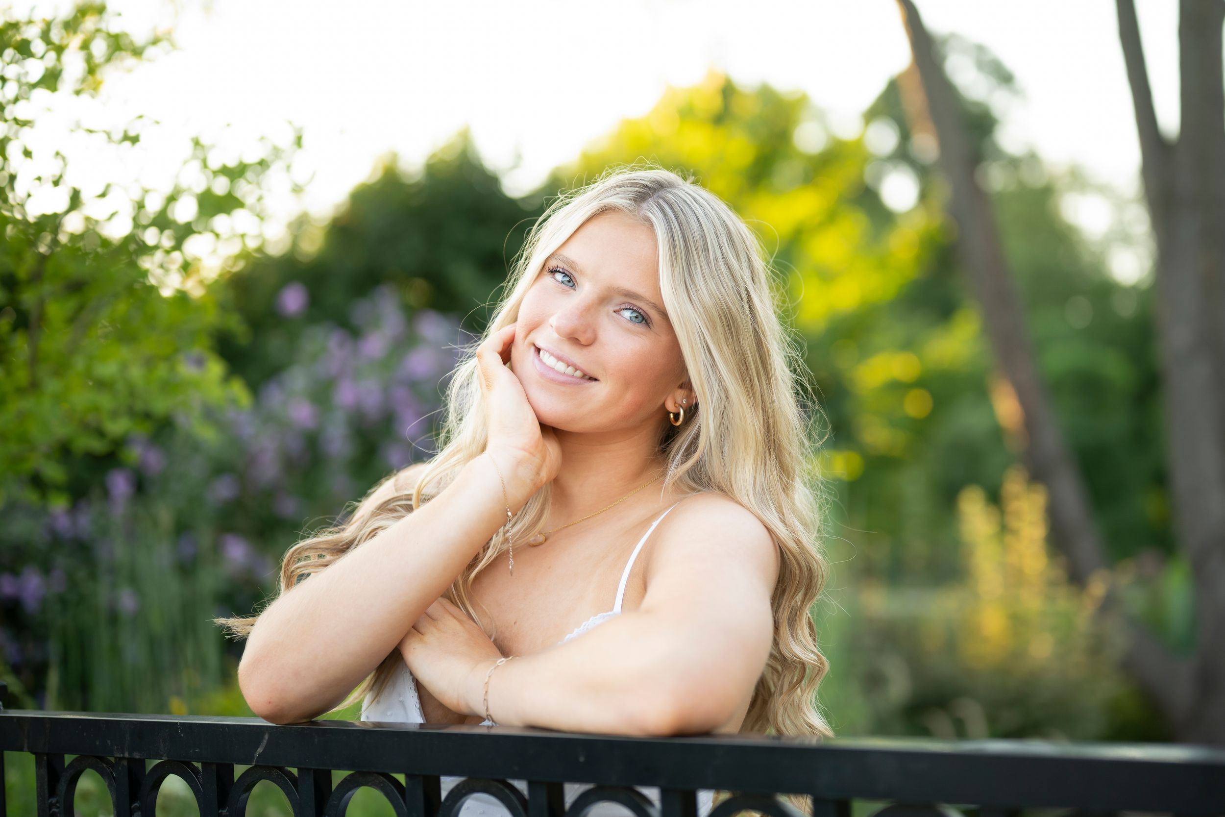 Smiling blonde woman leaning on iron fence in sunlit garden with purple flowers and green trees.
