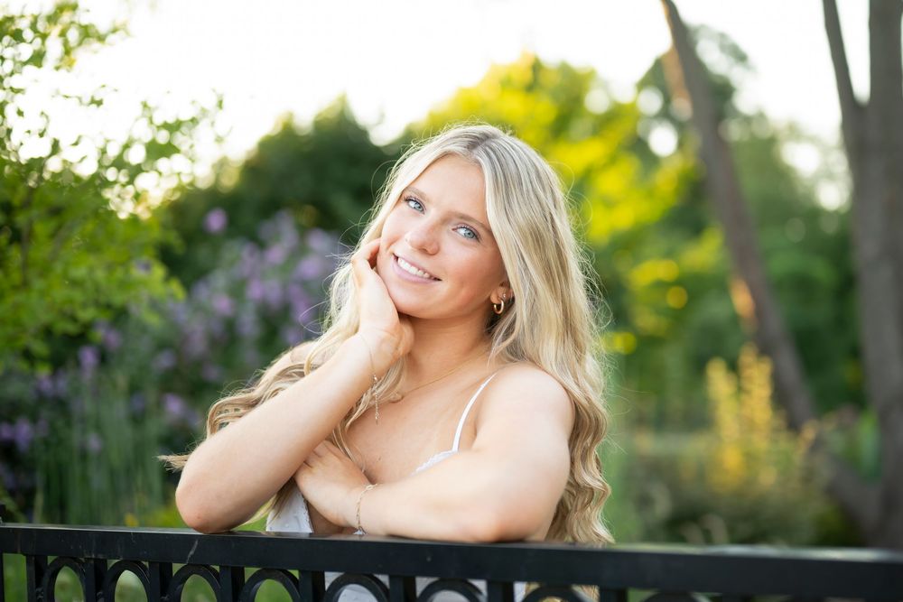 Smiling blonde woman leaning on iron fence in sunlit garden with purple flowers and green trees.
