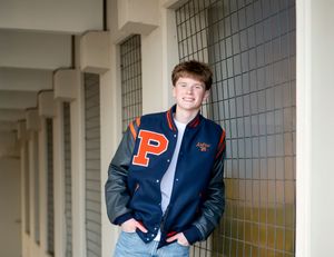 Teen boy smiling in navy varsity jacket with leather sleeves leaning against tiled wall.